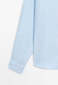 Chemise bleu clair à manches longues avec poignet boutonné, suspendue sur un fond blanc, montrant la texture du tissu et l'ourlet.