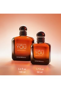 Dos frascos de perfume de vidrio ámbar con tapas plateadas redondeadas, etiquetados como "Stronger With You Absolutely" de Emporio Armani, mostrados en tamaños de 100 ml y 50 ml.