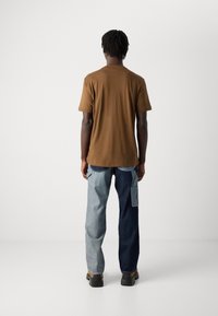 Mann med mørk hud og korte dreads som har på seg en brun t-skjorte og to-tone blå jeans, stående vendt mot en hvit bakgrunn.