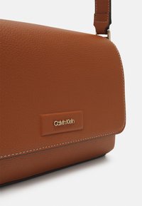 Brązowa torebka z teksturowanej skóry z klapą, białym przeszyciem i złotą plakietką z logo Calvin Klein na przodzie. Regulowany pasek przymocowany.