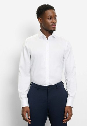 Mann trägt ein weißes, langärmeliges Hemd mit Knöpfen, das in dunkelblaue Hosen gesteckt ist, steht vor einem schlichten hellen Hintergrund und schaut seitlich.