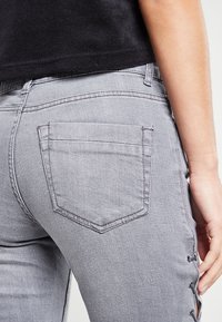 Šedé denimové džíny se přiléhavým střihem, zadními kapsami s zaobleným tvarem a bočními šněrovacími detaily podél nohy.
