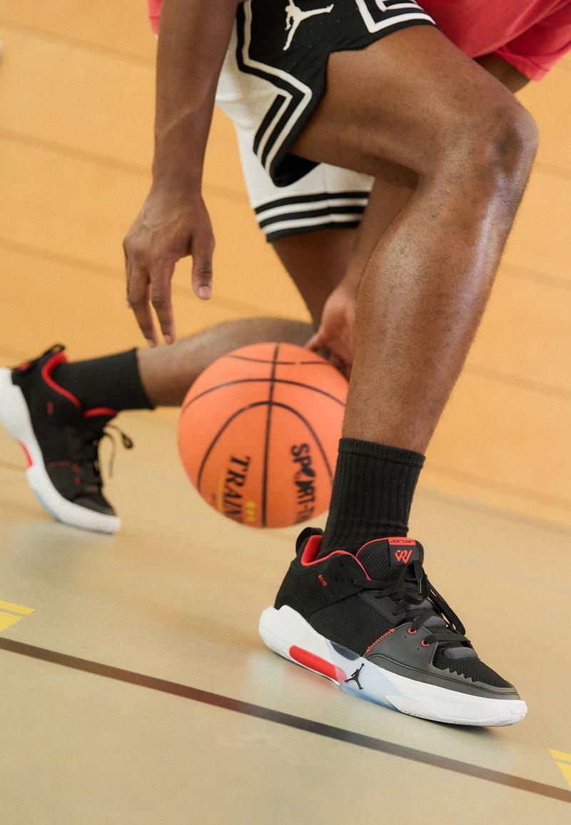 Černé sportovní boty s červenými a bílými prvky, s prodyšným svrškem, gumovou podrážkou a výrazným logem Jordan. Hrající basketbal.