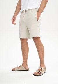 Beige katoenen shorts met een elastische tailleband en trekkoord, gecombineerd met lichtbruine sandalen met twee verstelbare banden.