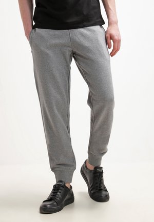Persoon die grijze joggingbroek en zwarte sneakers draagt, staat met handen in de zakken op een lichte achtergrond.