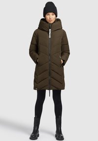 Olijfgroene pufferjas met chevronstiksels, hoge kraag, rits aan de voorkant en zijzakken. Draagt over zwarte leggings en enkelboots.