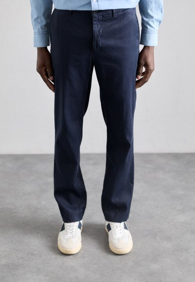Pantalons en lin bleu marine avec une coupe droite, dotés d'une fermeture éclair et d'un bouton à l'avant. Portés avec des chaussures blanches et des accents bleus.