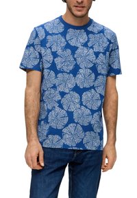 Niebieska koszulka z białym wzorem liści. Krótkie rękawy, okrągły dekolt, miękka faktura. Noszona z niebieskimi dżinsami.