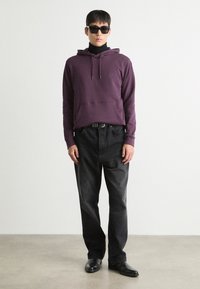 Fialová bavlnená mikina s predným vreckom, v kombinácii s čiernymi džínami. Čierne slnečné okuliare dokončujú outfit. Jednoduchý, moderný dizajn.