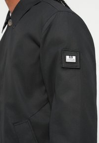 Giacca nera con una finitura liscia e opaca. Presenta un colletto a scollatura, chiusura a bottone e una patch con il marchio sulla manica sinistra.