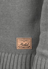 Maglione grigio lavorato a maglia con orlo a coste. Presenta un'etichetta rettangolare beige con il testo "Solid United Traditions" e elementi decorativi.