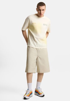 Młody mężczyzna stoi ze złożonymi za plecami rękami, ubrany w kremową koszulkę, beżowe szorty do kolan, białe skarpetki i kolorowe trampki.