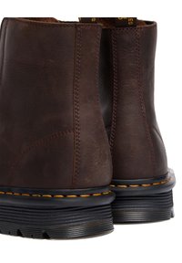 Paire de bottes en cuir marron foncé avec semelles épaisses noires à rainures et coutures jaunes le long du bord de la semelle, vue de dos.