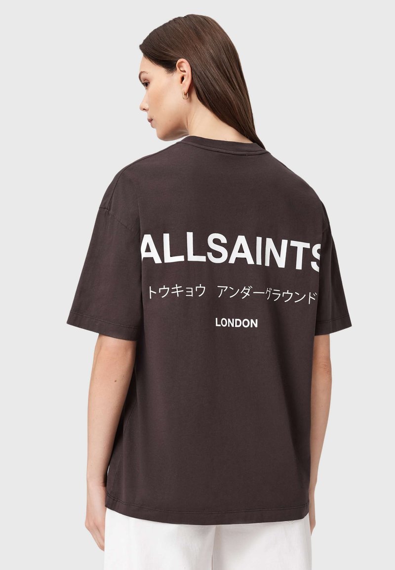 Žena s dlhými hnedými vlasmi nosiaca čierne oversized tričko s nápisom "ALLSAINTS", japonským textom a "LONDON" vytlačeným na chrbte.