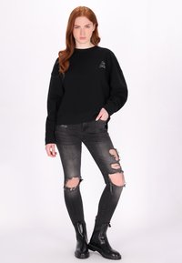 Černá mikina s kulatým výstřihem, oversized střih, v kombinaci se zvlněnými černými džíny s roztrhanými koleny a černými kotníkovými botami.