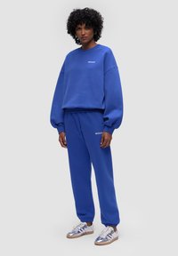 Niebieska bluza i dopasowane spodnie dresowe z miękkiego materiału; szerokie rękawy i elastyczne mankiety; biały logo na klatce piersiowej i spodniach.