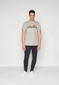 Man standing wearing a grey ellesse t-shirt, black joggers, and white sneakers against a plain light background.