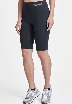 Femme portant un short de sport noir taille haute jusqu'aux genoux avec le logo "hummel", associé à des chaussures de course blanches et grises sur fond blanc.