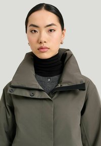 Oliven grøn vandtæt jakke med høj krave, sorte accenter og trykknaplukninger, lagdelt over en grå turtleneck sweater.