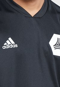 Jersey atlético negro hecho de tela texturizada, con el logo blanco de Adidas y un parche hexagonal blanco con un diseño negro en el pecho.