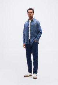 Πουκάμισο από τζιν με κουμπιά και μία τσέπη στο στήθος, συνδυασμένο με σκούρα μπλε τζιν και λευκά αθλητικά παπούτσια. Το πουκάμισο έχει χαλαρή εφαρμογή και γιακά.