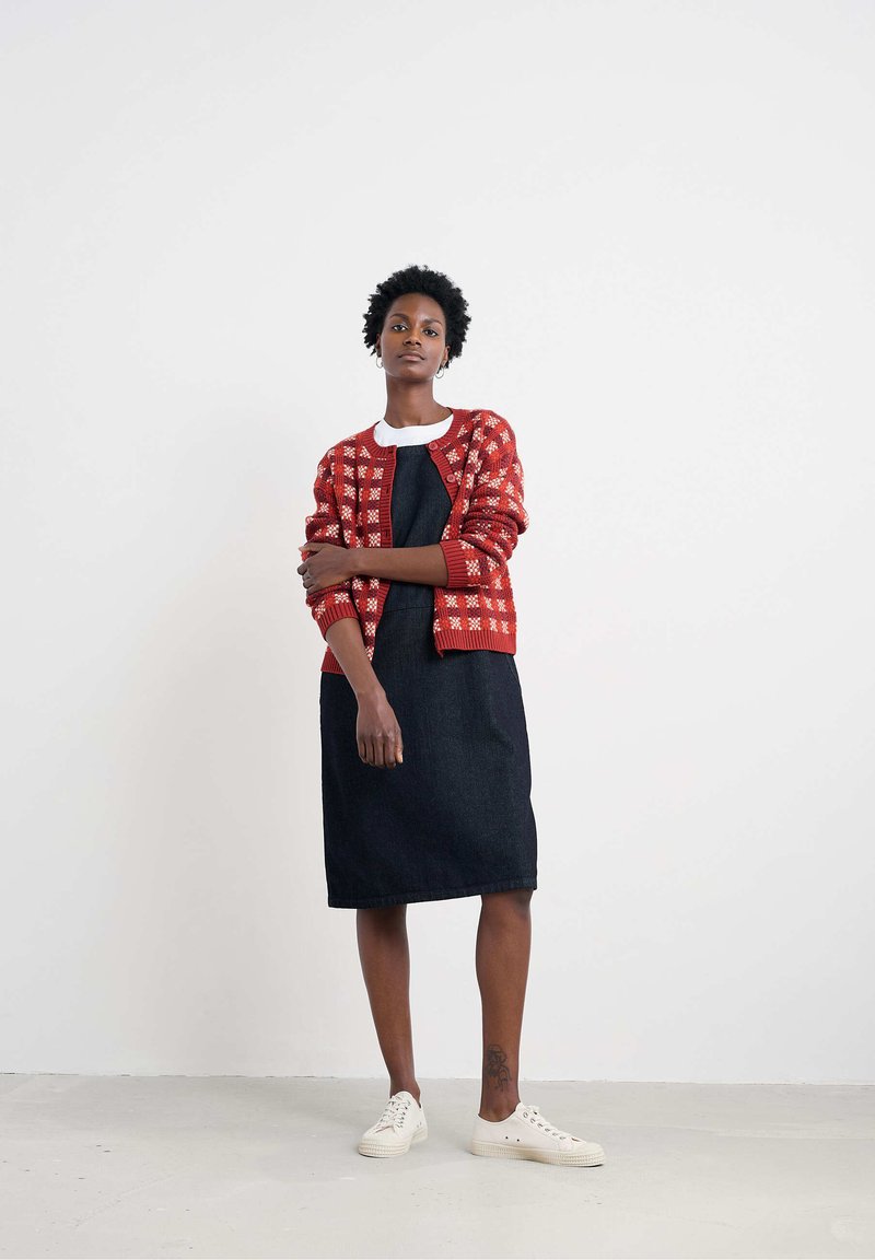 Žena stojaca proti bielej stene, oblečená v kardigáne s červeným vzorom, čiernych šatách, bielych teniskách a kruhových náušniciach, so viditeľným tetovaním na členku.