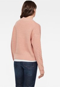 Ljusrosa stickad tröja med rundad halsringning, strukturerat mönster och ribbade ärmslut. Utförd över mörkblå jeans, sedd bakifrån.