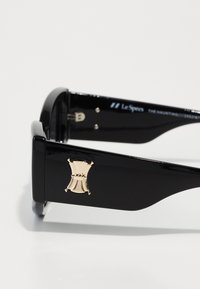 Svarte solbriller med blank finish, med et gullsymbol p�å armen og et elegant, vinklet design. Metallkomponenter er synlige.