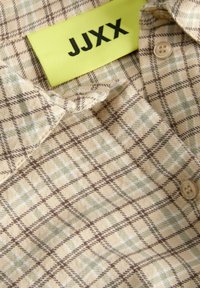 Chemise à carreaux beige et marron avec des boutons et une étiquette jaune indiquant un texte noir "JJXX" à l’intérieur du col.