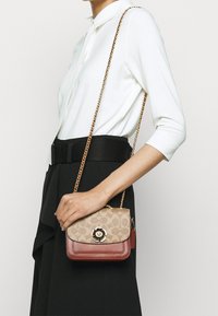 Donna in top bianco e gonna nera che tiene una borsa crossbody beige e marrone con chiusura floreale e tracolla dorata.