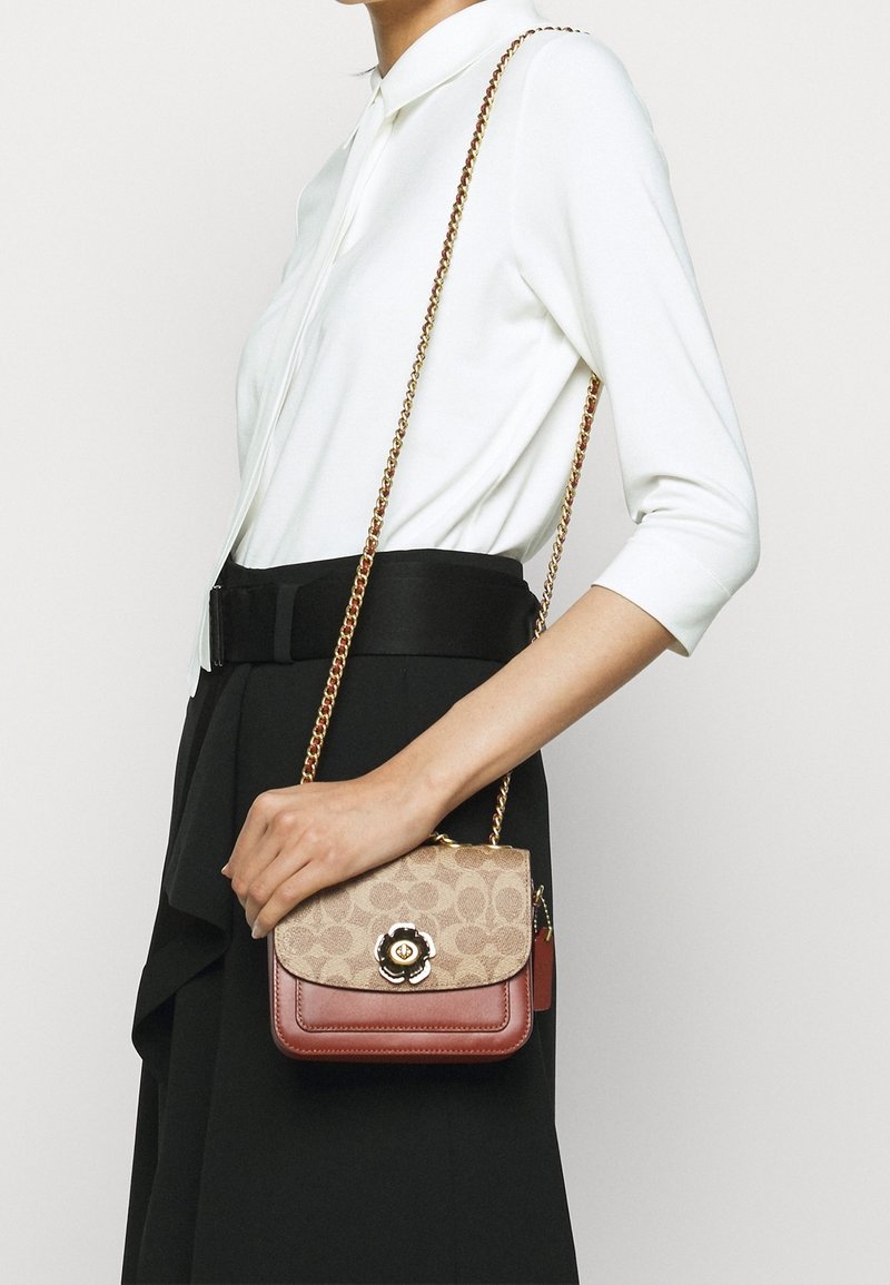 Donna in top bianco e gonna nera che tiene una borsa crossbody beige e marrone con chiusura floreale e tracolla dorata.