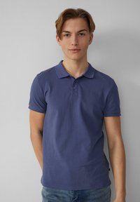 Koszulka polo z krótkim rękawem w głębokim fioletowym odcieniu, z kołnierzykiem, trzema guzikami oraz miękkim, teksturowanym materiałem. Noszona z niebieskimi dżinsami.