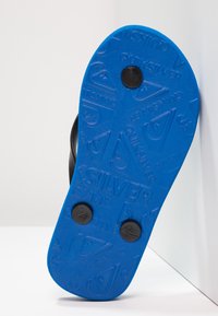 Czarna sandał z niebieską gumową podeszwą, z teksturowanymi wzorami zapewniającymi uchwyt i dwoma czarnymi okrągłymi akcentami. Podeszwa zawiera szczegóły dotyczące marki.