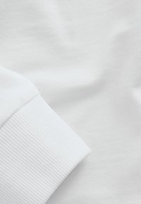 Biely textil zblízka, ktorý zobrazuje rebrovaný manžet a hladký povrch. Materiál sa zdá byť mäkký s jednoduchým, bez vzorovým povrchom.