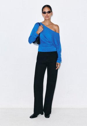Femme portant un top bleu épaules dénudées, un pantalon noir à jambes larges, des chaussures noires, des lunettes de soleil foncées, et un petit sac à bandoulière noir.