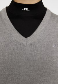 Harmaa neulepaita V-pääntiellä, jossa on hienovarainen tekstuuri ja pieni logo rinnassa, kerroksittain mustan poolopaidan päällä.