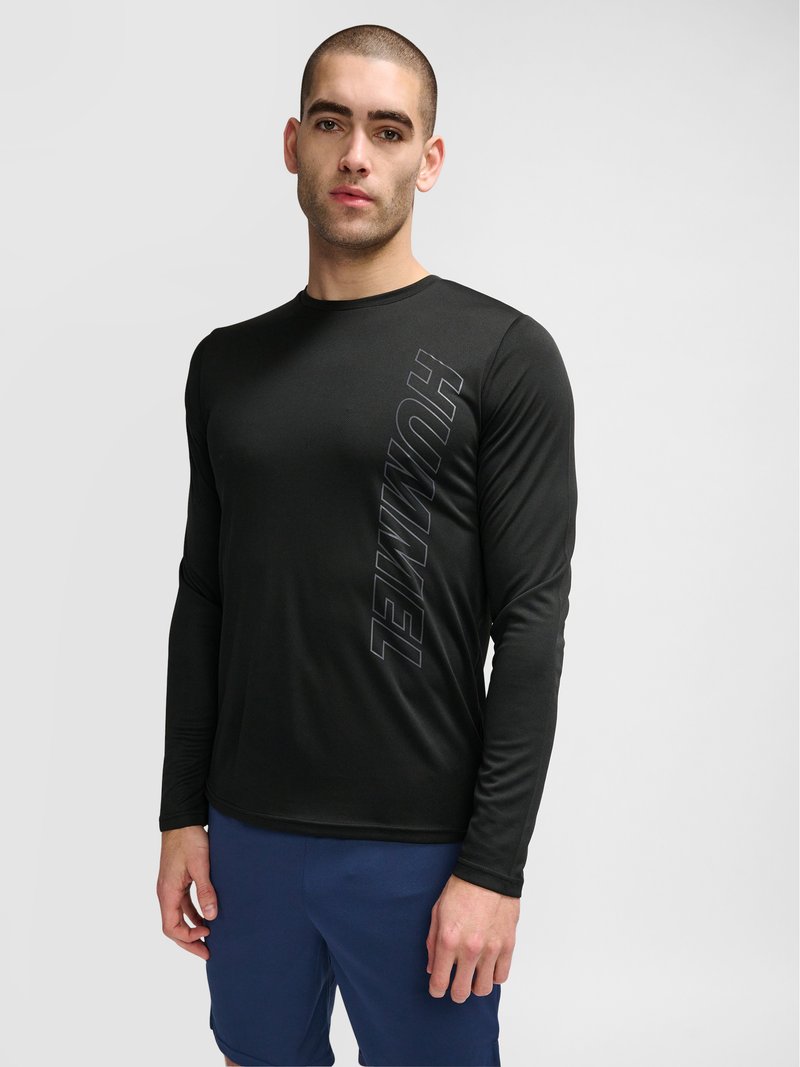 Homme aux cheveux courts portant un t-shirt noir à manches longues avec « HUMMEL » imprimé verticalement sur le côté gauche, associé à un short bleu, debout devant un fond gris.