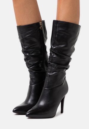 Mustat nahkaiset polvipituiset saappaat, joissa on terävä kärki, reno tekstuuri ja kultainen vetoketju sivussa. Korkeat stiletto-korot.