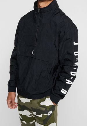 Person trägt eine schwarze Jordan Half-Zip-Jacke mit weißem Logo und Text am Ärmel, kombiniert mit grünen und weißen Nike Camouflage-Hosen.