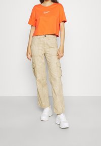 Oranget cropped t-shirt med vit logotyp, parat med beige cargobyxor med ett virvlande mönster och vita snörskor.