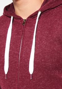 Bordeauxrode zip-up hoodie met witte trekkoorden, uitgevoerd in een gestructureerde stof en metalen ritssluiting. Close-up van de halslijn en voorkant.