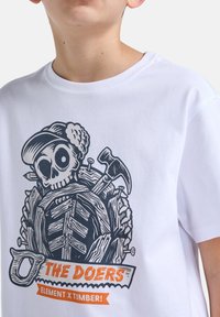 Weißes T-Shirt mit kurzen Ärmeln, das ein schwarzes und oranges grafisches Design eines Skeletts mit Werkzeugen zeigt, beschriftet mit "DIE TÄTER" am unteren Rand.