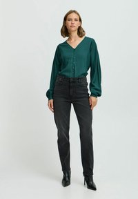 Blusa teal a maniche lunghe a coste con scollo a V e chiusura con bottoni, abbinata a jeans neri a vita alta e scarpe con tacco nere.