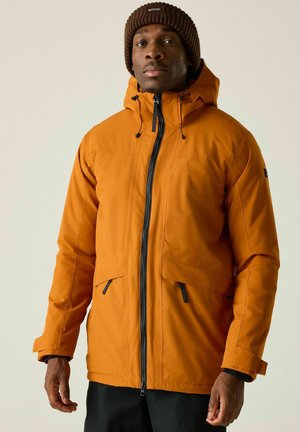 Mężczyzna w brązowej dzianinowej czapce i pomarańczowej kurtce z kapturem, z czarnym zamkiem błyskawicznym i kieszeniami, stojący na tle jasnego, jednolitego tła.