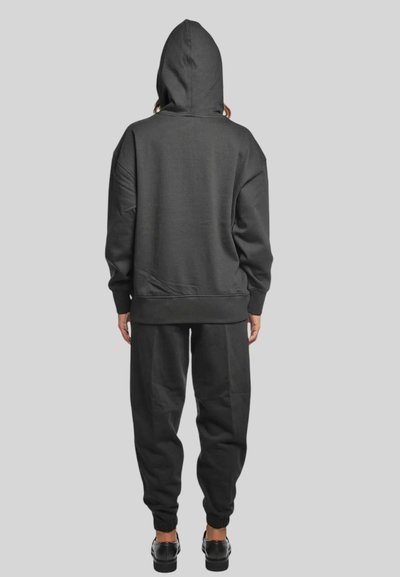 Persona che indossa una felpa con cappuccio nera e pantaloni della tuta neri abbinati, in piedi di spalle su uno sfondo grigio chiaro uniforme.