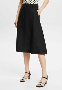 Falda A-line de lino negra con cinturilla y bolsillos funcionales, combinada con sandalias de tiras blanco y negro con tacón contundente.