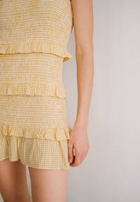 Žlutý dvoudílný set se smockovaným topem s volány a odpovídající sukní s kostkovaným vzorem, zvýrazňující texturu a designové detaily.