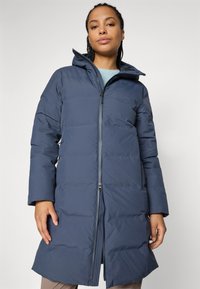 Vrouw in een lange, navyblauwe pufferjas met hoge kraag, staand tegen een effen lichte achtergrond.