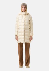 Ocieplana kremowa kurtka z wysokim kołnierzem i regulowanym kapturem, z bocznymi kieszeniami, w zestawie z brązowymi spodniami i wzorzystą czapką dzianinową.