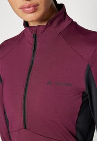 Haut de sport bordeaux avec un col haut, des accents noirs sur les côtés et une demi-fermeture éclair. Présente le logo noir VAUDE au niveau de la poitrine.